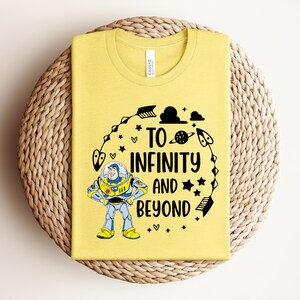 Peut inclure: T-shirt jaune avec un graphisme de Buzz l'&Eacute;clair et l'inscription "To Infinity and Beyond". Le motif comprend des &eacute;toiles, des plan&egrave;tes et des fl&egrave;ches. Le t-shirt est pli&eacute; sur un dessous de plat circulaire tiss&eacute;.