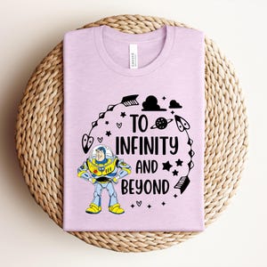 Peut inclure: Un t-shirt lilas avec un graphisme de Buzz l'&Eacute;clair et l'inscription "To Infinity and Beyond" en noir. Le motif comprend des &eacute;toiles, des plan&egrave;tes et d'autres &eacute;l&eacute;ments sur le th&egrave;me de l'espace. Le t-shirt est pli&eacute; sur un tapis tiss&eacute;.