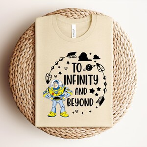 Peut inclure: T-shirt beige avec un graphisme color&eacute; de Buzz l'&Eacute;clair et le texte "To Infinity and Beyond". Le motif comprend des &eacute;toiles, des fl&egrave;ches et des plan&egrave;tes. Le t-shirt est pr&eacute;sent&eacute; sur une surface circulaire tiss&eacute;e.