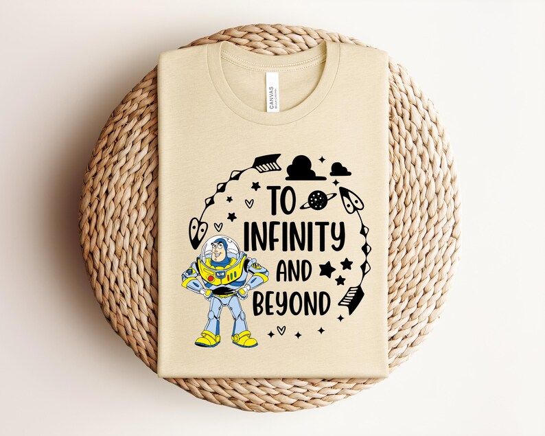 Peut inclure: T-shirt beige avec un graphisme color&eacute; de Buzz l'&Eacute;clair et le texte "To Infinity and Beyond". Le motif comprend des &eacute;toiles, des fl&egrave;ches et des plan&egrave;tes. Le t-shirt est pr&eacute;sent&eacute; sur une surface circulaire tiss&eacute;e.