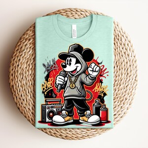 May include: A light green t-shirt featuring a cartoon Mickey Mouse character in a hip-hop style outfit, holding a microphone. The design includes graffiti art, a boombox, and spray paint cans. The shirt is folded on a woven, circular surface.
