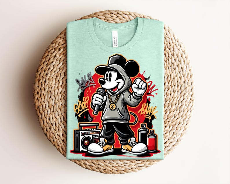 May include: A light green t-shirt featuring a cartoon Mickey Mouse character in a hip-hop style outfit, holding a microphone. The design includes graffiti art, a boombox, and spray paint cans. The shirt is folded on a woven, circular surface.
