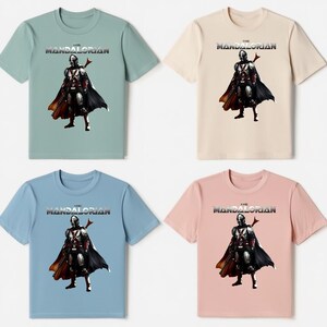 Op de afbeelding: Vier T-shirts in verschillende kleuren: lichtgroen, beige, lichtblauw en roze. Elk shirt heeft een afbeelding van een personage uit "The Mandalorian" met de titel van de show erboven. Het personage draagt een zwarte cape.