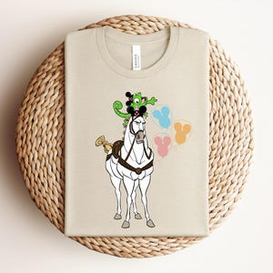 May include: A beige t-shirt featuring a cartoon illustration of a white horse with a black harness and a gold bow. The horse has a green chameleon and black Mickey Mouse ears on its head. Three colorful balloons are to the right.
