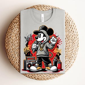 May include: A light gray t-shirt featuring a cartoon Mickey Mouse in a hip-hop style. Mickey is wearing a gray hoodie, black pants, and a gold chain, holding a microphone. Graffiti art and a boombox are also visible.