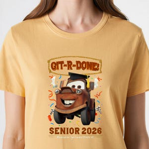 Op de afbeelding: Mosterdgeel T-shirt met een cartoonafbeelding van Takel uit de film Cars, met een afstudeerhoed. De tekst "GIT-R-DONE!" staat boven Takel en "SENIOR 2026" staat eronder. Radiator Springs Class of is ook gedrukt.