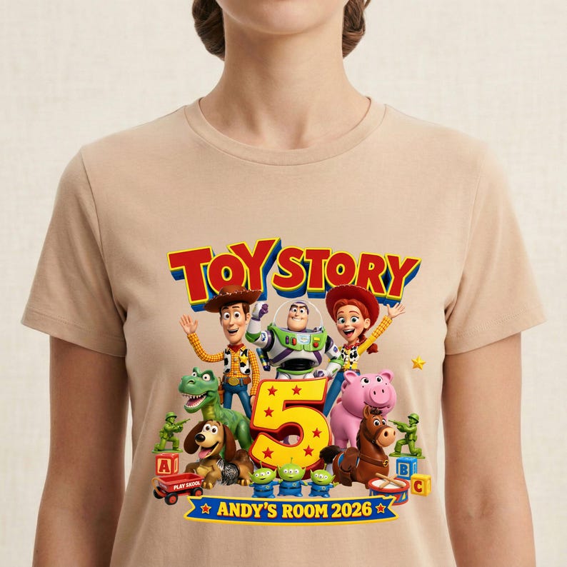 May include: A beige t-shirt featuring a colorful Toy Story graphic with Woody, Buzz Lightyear, Jessie, Rex, Slinky Dog, Hamm, Bullseye, and green army men. The design includes the text Toy Story, a large number 5, and Andy's Room 2026.