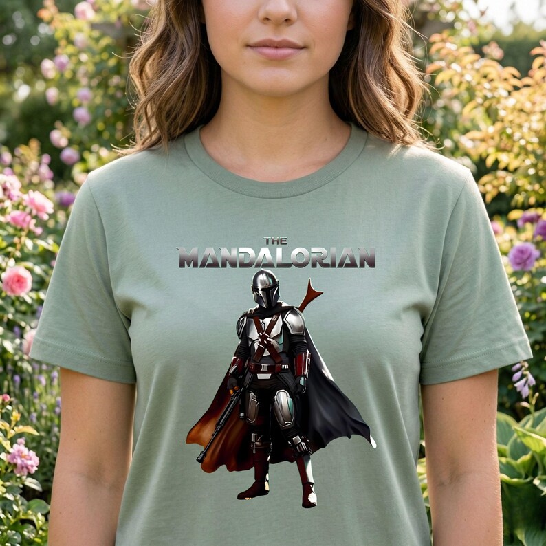 Op de afbeelding: Saliegroen T-shirt met de tekst "THE MANDALORIAN" en een afbeelding van het Mandalorian-personage in harnas, cape en wapen. Het ontwerp is in zilver, zwart, rood en bruin.
