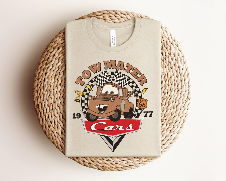 Puede incluir: Una camiseta beige con un estampado gr&aacute;fico del personaje Tow Mater de la pel&iacute;cula Cars. El dise&ntilde;o incluye una bandera a cuadros, un trofeo dorado, una se&ntilde;al de la Ruta 66, rayos y el texto Tow Mater, 19, 77 y Cars en color rojo.