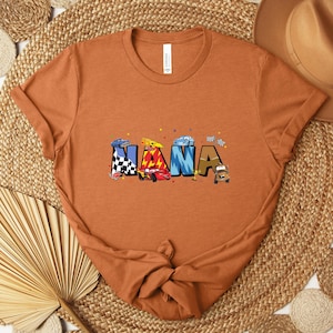K&ouml;nnte beinhalten: Ein orangefarbenes T-Shirt mit dem Wort "NANA" in bunten Buchstaben im Auto-Design. Die Buchstaben sind mit Rennwagen und Zielflaggen verziert. Das T-Shirt ist am unteren Ende geknotet und wird auf einer gewebten Matte pr&auml;sentiert.
