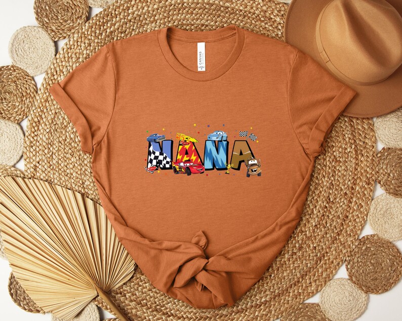 K&ouml;nnte beinhalten: Ein orangefarbenes T-Shirt mit dem Wort "NANA" in bunten Buchstaben im Auto-Design. Die Buchstaben sind mit Rennwagen und Zielflaggen verziert. Das T-Shirt ist am unteren Ende geknotet und wird auf einer gewebten Matte pr&auml;sentiert.