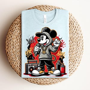 May include: Light blue t-shirt featuring a cartoon Mickey Mouse character in a hip-hop style. Mickey is wearing a hoodie, cap, and gold chain, holding a microphone. Graffiti art and a boombox are also visible.