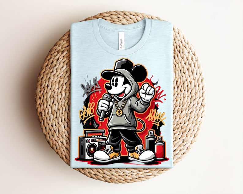 May include: Light blue t-shirt featuring a cartoon Mickey Mouse character in a hip-hop style. Mickey is wearing a hoodie, cap, and gold chain, holding a microphone. Graffiti art and a boombox are also visible.