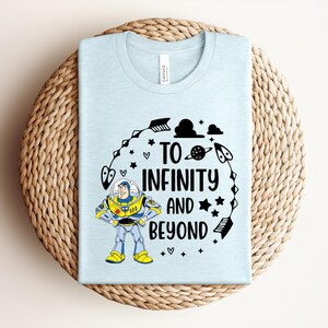 Peut inclure: Un t-shirt bleu clair avec l'inscription noire "TO INFINITY AND BEYOND", accompagn&eacute; d'un dessin anim&eacute; de Buzz l'&Eacute;clair. Le motif comprend des &eacute;toiles, des fl&egrave;ches et une plan&egrave;te. Le t-shirt est pli&eacute; sur un tapis circulaire tiss&eacute;.