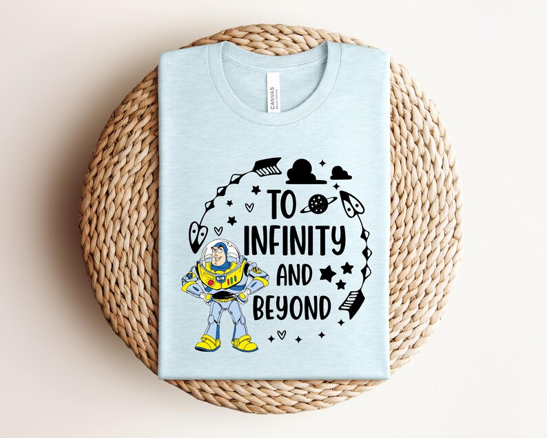 Peut inclure: Un t-shirt bleu clair avec l'inscription noire "TO INFINITY AND BEYOND", accompagn&eacute; d'un dessin anim&eacute; de Buzz l'&Eacute;clair. Le motif comprend des &eacute;toiles, des fl&egrave;ches et une plan&egrave;te. Le t-shirt est pli&eacute; sur un tapis circulaire tiss&eacute;.