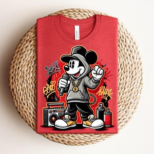 May include: Red t-shirt featuring a cartoon Mickey Mouse in a hip-hop style, wearing a hoodie, gold chain, and holding a microphone. The design includes graffiti art, a boombox, and spray paint cans. The shirt is folded on a woven surface.