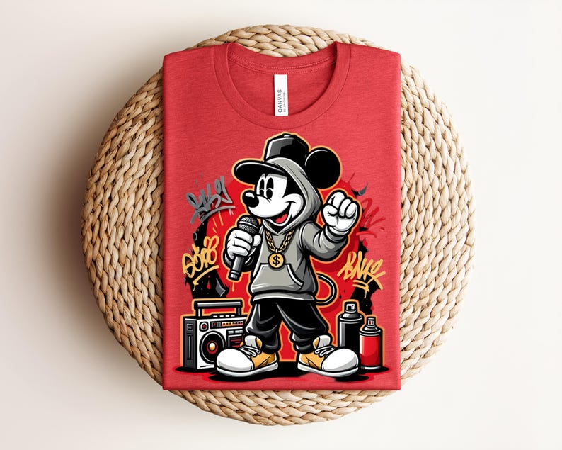 May include: Red t-shirt featuring a cartoon Mickey Mouse in a hip-hop style, wearing a hoodie, gold chain, and holding a microphone. The design includes graffiti art, a boombox, and spray paint cans. The shirt is folded on a woven surface.