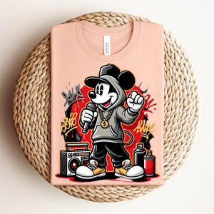 May include: Peach-colored t-shirt featuring a cartoon Mickey Mouse in a hip-hop style. Mickey is wearing a gray hoodie, black pants, and a gold chain, holding a microphone. The design includes graffiti art and a boombox.