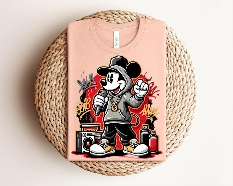 May include: Peach-colored t-shirt featuring a cartoon Mickey Mouse in a hip-hop style. Mickey is wearing a gray hoodie, black pants, and a gold chain, holding a microphone. The design includes graffiti art and a boombox.