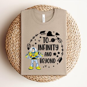 Peut inclure: Un t-shirt beige avec un motif color&eacute; de Buzz l'&Eacute;clair et l'inscription noire "TO INFINITY AND BEYOND". Le motif comprend des &eacute;toiles, des plan&egrave;tes et d'autres &eacute;l&eacute;ments sur le th&egrave;me de l'espace. Le t-shirt est pli&eacute; sur un tapis circulaire tiss&eacute;.