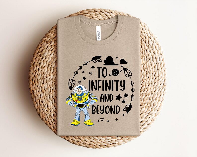 Peut inclure: Un t-shirt beige avec un motif color&eacute; de Buzz l'&Eacute;clair et l'inscription noire "TO INFINITY AND BEYOND". Le motif comprend des &eacute;toiles, des plan&egrave;tes et d'autres &eacute;l&eacute;ments sur le th&egrave;me de l'espace. Le t-shirt est pli&eacute; sur un tapis circulaire tiss&eacute;.