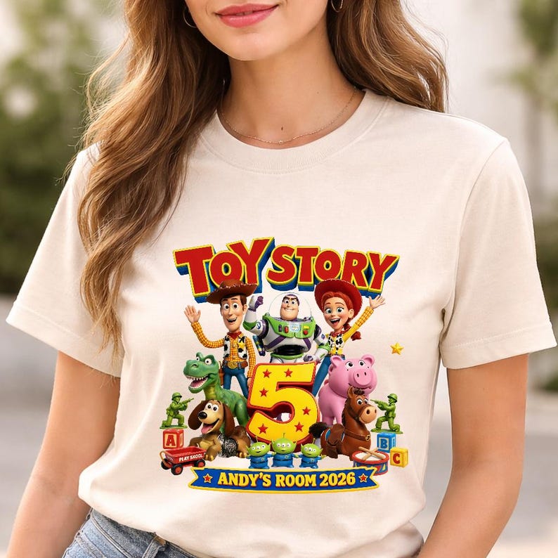 May include: A cream-colored short-sleeve t-shirt featuring a colorful graphic of Toy Story characters including Woody, Buzz Lightyear, Jessie, Rex, Slinky Dog, Hamm, Bullseye, and green army men surrounding a large yellow number 5 and text.