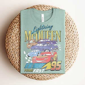 Puede incluir: Una camiseta verde claro con un gr&aacute;fico de Rayo McQueen y otros personajes de Cars. El dise&ntilde;o incluye el texto "Lightning McQueen", "95" y "Think Fast" en un estilo vintage. La camiseta est&aacute; doblada sobre una estera tejida.