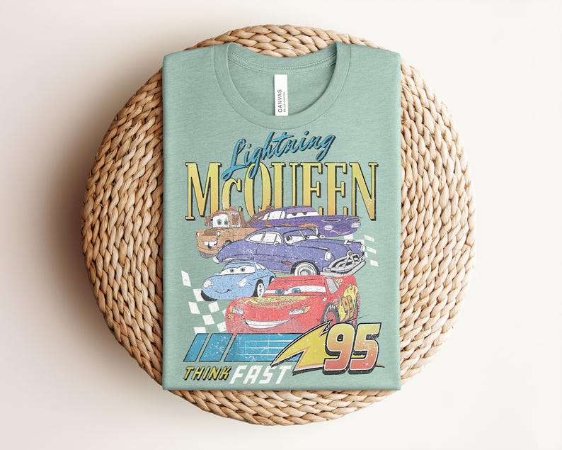 Puede incluir: Una camiseta verde claro con un gr&aacute;fico de Rayo McQueen y otros personajes de Cars. El dise&ntilde;o incluye el texto "Lightning McQueen", "95" y "Think Fast" en un estilo vintage. La camiseta est&aacute; doblada sobre una estera tejida.