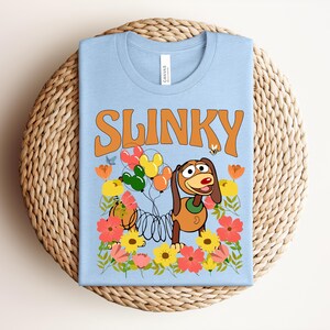 K&ouml;nnte beinhalten: Hellblaues T-Shirt mit dem Wort "SLINKY" in orangefarbenen Blockbuchstaben. Das Shirt hat eine Grafik von Slinky Dog mit Luftballons und Blumen. Das T-Shirt ist auf einer gewebten, kreisf&ouml;rmigen Oberfl&auml;che gefaltet.