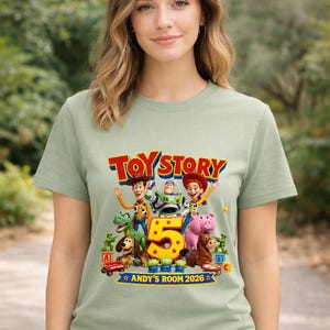 May include: A light sage green t-shirt featuring a colorful graphic of Toy Story characters including Woody, Buzz Lightyear, Jessie, Rex, Slinky Dog, Hamm, and Bullseye. The design includes a large yellow number 5 and the text TOY STORY ANDY'S ROOM 2026.