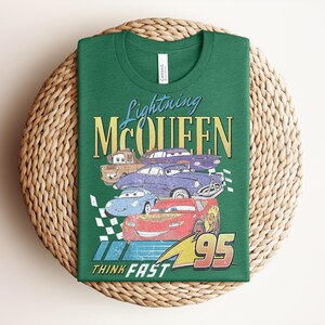 Puede incluir: Camiseta verde con un dise&ntilde;o de Rayo McQueen y otros personajes de Cars. El dise&ntilde;o incluye el texto "Lightning McQueen", "95" y "Think Fast". La camiseta est&aacute; doblada sobre una estera tejida.