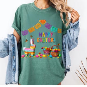 K&ouml;nnte beinhalten: T&uuml;rkisgr&uuml;nes T-Shirt mit einer farbenfrohen Ostergrafik. Das Design umfasst eine Pi&ntilde;ata, Osterk&ouml;rbe, Schmetterlinge und den Text "HAPPY EASTER!" Das Shirt hat hochgekrempelte &Auml;rmel und eine lockere Passform.