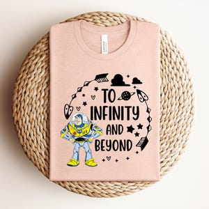 Peut inclure: Un t-shirt rose clair avec un dessin de Buzz l'&Eacute;clair et l'inscription "To Infinity and Beyond" en noir. Le motif comprend des &eacute;toiles, des fl&egrave;ches et une plan&egrave;te. Le t-shirt est pli&eacute; sur un dessous de plat circulaire tiss&eacute;.