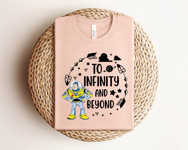 Peut inclure: Un t-shirt rose clair avec un dessin de Buzz l'&Eacute;clair et l'inscription "To Infinity and Beyond" en noir. Le motif comprend des &eacute;toiles, des fl&egrave;ches et une plan&egrave;te. Le t-shirt est pli&eacute; sur un dessous de plat circulaire tiss&eacute;.