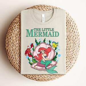 May include: A light beige t-shirt featuring a graphic of the character Ariel from The Little Mermaid sitting in a pink seashell. The design includes a red crab, a yellow fish, green seaweed, and blue bubbles, with the text The Little Mermaid above.