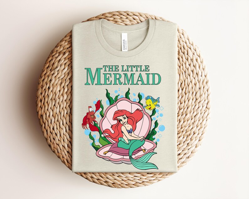 May include: A light beige t-shirt featuring a graphic of the character Ariel from The Little Mermaid sitting in a pink seashell. The design includes a red crab, a yellow fish, green seaweed, and blue bubbles, with the text The Little Mermaid above.