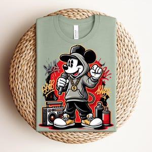 May include: Sage green t-shirt featuring a cartoon Mickey Mouse character in a hip-hop style outfit, holding a microphone. The design includes graffiti art, a boombox, and spray paint cans. The shirt is displayed on a woven, circular placemat.