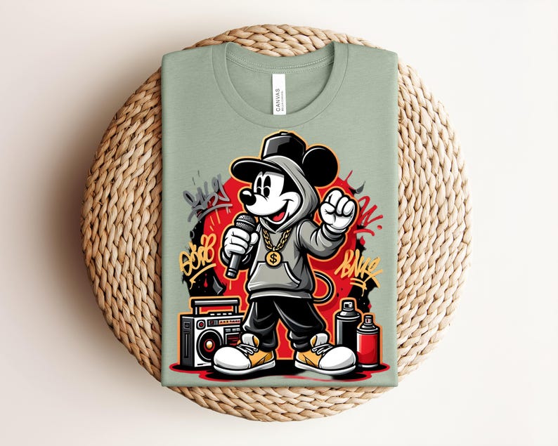 May include: Sage green t-shirt featuring a cartoon Mickey Mouse character in a hip-hop style outfit, holding a microphone. The design includes graffiti art, a boombox, and spray paint cans. The shirt is displayed on a woven, circular placemat.