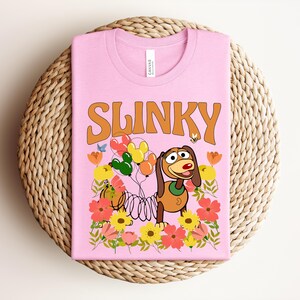 K&ouml;nnte beinhalten: Rosafarbenes T-Shirt mit dem Wort "SLINKY" in Orange. Das Design zeigt Slinky Dog, Ballons, Blumen und eine Biene. Das T-Shirt liegt auf einem runden Untersetzer aus Korbgeflecht.