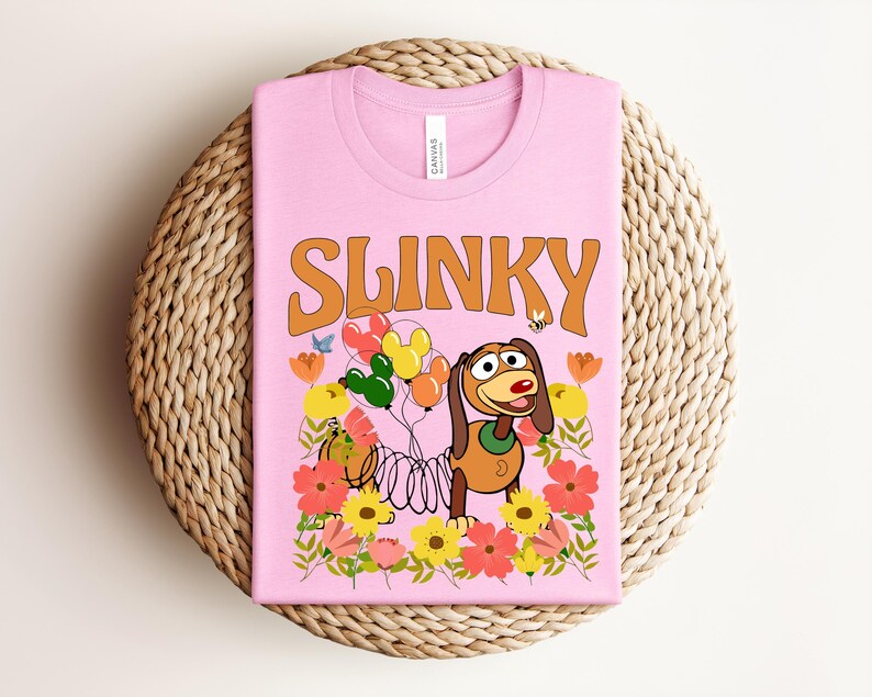 K&ouml;nnte beinhalten: Rosafarbenes T-Shirt mit dem Wort "SLINKY" in Orange. Das Design zeigt Slinky Dog, Ballons, Blumen und eine Biene. Das T-Shirt liegt auf einem runden Untersetzer aus Korbgeflecht.