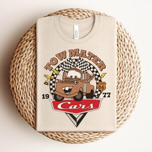 Puede incluir: Una camiseta de color crema con un gr&aacute;fico del personaje Tow Mater de la pel&iacute;cula Cars. El dise&ntilde;o incluye una bandera a cuadros, un trofeo dorado, una se&ntilde;al de la Ruta 66, el texto Tow Mater, 1977 y el logotipo rojo de Cars en la parte inferior.