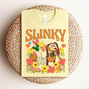 K&ouml;nnte beinhalten: Ein hellgelbes T-Shirt mit dem Wort "SLINKY" in orangefarbener Retro-Schrift. Das T-Shirt zeigt einen Zeichentrickhund, der Luftballons h&auml;lt, umgeben von Blumen. Das T-Shirt ist auf einer gewebten, kreisf&ouml;rmigen Matte gefaltet.