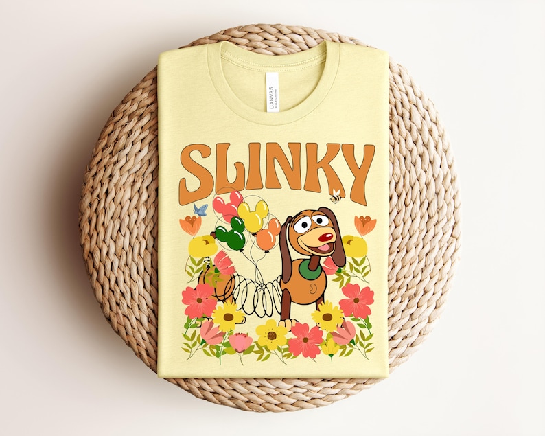 K&ouml;nnte beinhalten: Ein hellgelbes T-Shirt mit dem Wort "SLINKY" in orangefarbener Retro-Schrift. Das T-Shirt zeigt einen Zeichentrickhund, der Luftballons h&auml;lt, umgeben von Blumen. Das T-Shirt ist auf einer gewebten, kreisf&ouml;rmigen Matte gefaltet.