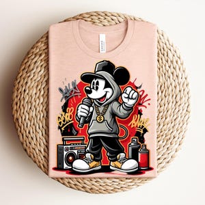 May include: A light pink t-shirt featuring a cartoon Mickey Mouse character in a hip-hop style. Mickey is wearing a gray hoodie, black pants, and a gold chain, holding a microphone. Graffiti art and a boombox are also visible.