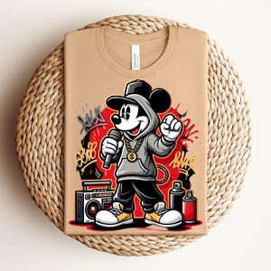 May include: Tan t-shirt featuring a cartoon Mickey Mouse character in a hip-hop style. Mickey is wearing a gray hoodie, gold chain, and black pants, holding a microphone. Graffiti art and a boombox are also visible.