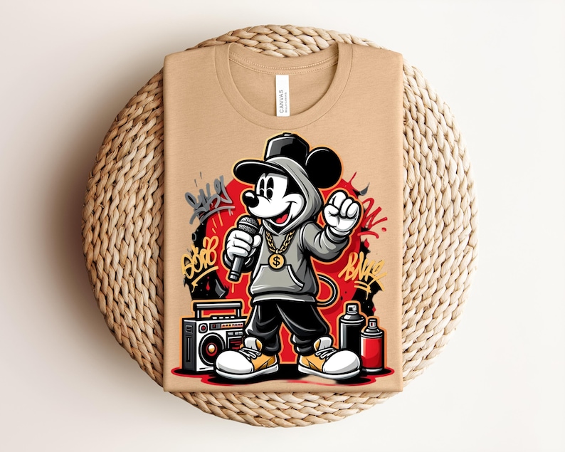 May include: Tan t-shirt featuring a cartoon Mickey Mouse character in a hip-hop style. Mickey is wearing a gray hoodie, gold chain, and black pants, holding a microphone. Graffiti art and a boombox are also visible.