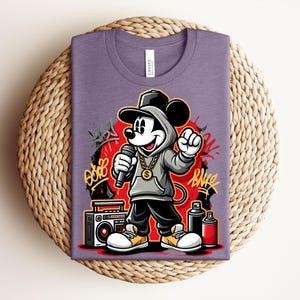 May include: Purple t-shirt featuring a cartoon Mickey Mouse in a hip-hop style outfit, holding a microphone. The design includes graffiti art, a boombox, and spray paint cans. The shirt is folded on a woven surface.