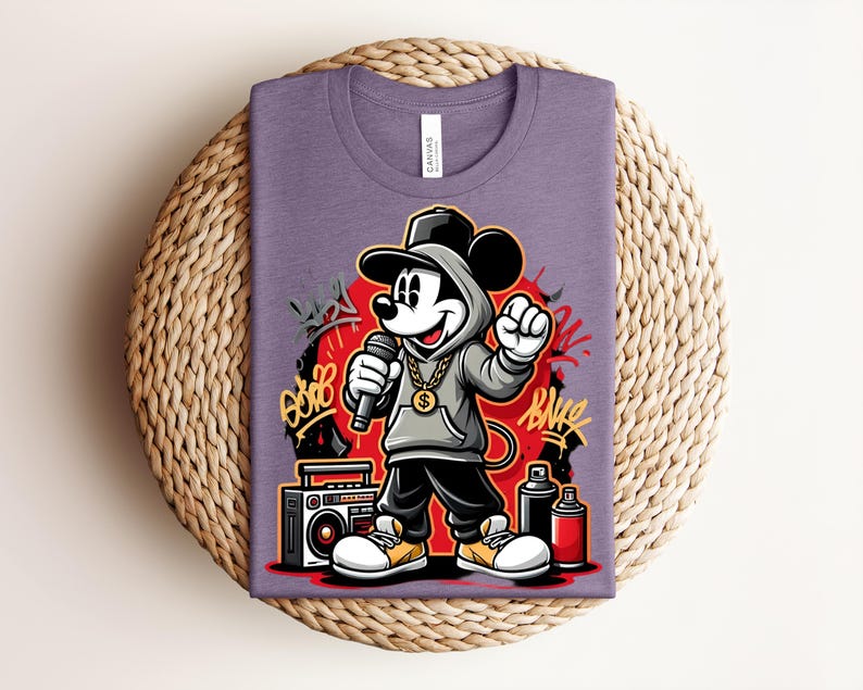 May include: Purple t-shirt featuring a cartoon Mickey Mouse in a hip-hop style outfit, holding a microphone. The design includes graffiti art, a boombox, and spray paint cans. The shirt is folded on a woven surface.
