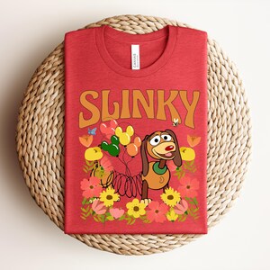 K&ouml;nnte beinhalten: Rotes T-Shirt mit dem Wort "SLINKY" in orangefarbenen Buchstaben. Das Shirt zeigt ein Cartoon-Bild von Slinky Dog mit Luftballons und Blumen. Das Shirt ist auf einer gewebten, kreisf&ouml;rmigen Matte gefaltet.