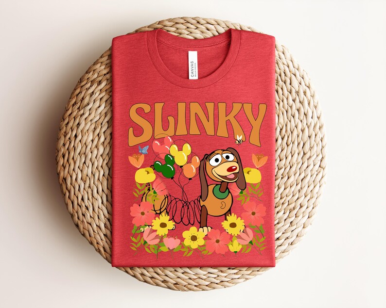 K&ouml;nnte beinhalten: Rotes T-Shirt mit dem Wort "SLINKY" in orangefarbenen Buchstaben. Das Shirt zeigt ein Cartoon-Bild von Slinky Dog mit Luftballons und Blumen. Das Shirt ist auf einer gewebten, kreisf&ouml;rmigen Matte gefaltet.
